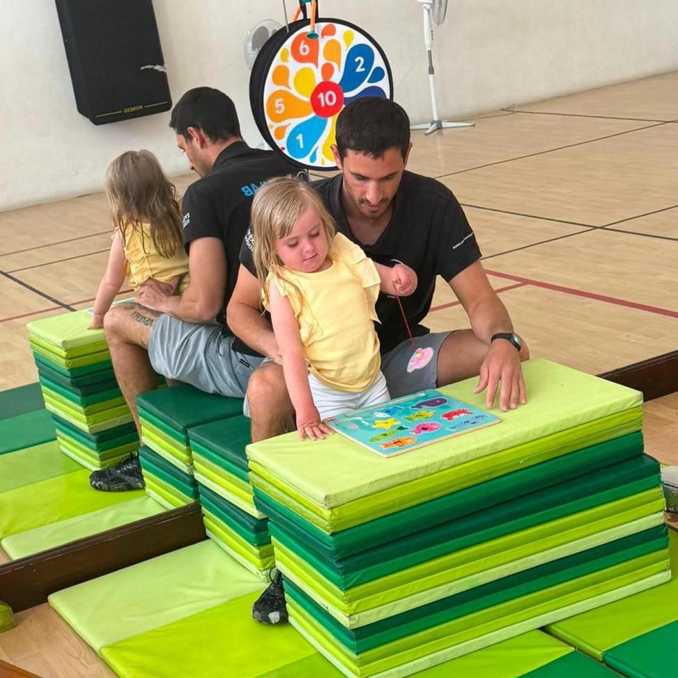 A young blonde girl wearing a yellow t short and white shorts. She is leant up against a stack of green play mats, playing with a coordination jigsaw game in front of her. She is being supported by a man with brown haor, wearing a black polo short and grey shorts. He is sat being her, on a another stack of green mats. Behind them both is a large mirrored wall.