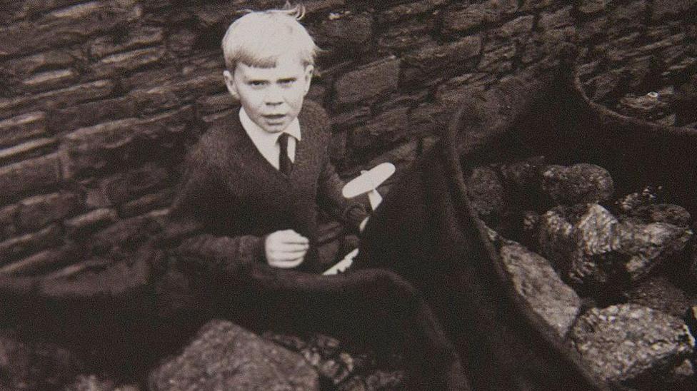 A black and white image of an eight year old boy. His hair is white and he is holding a toy aeroplane, while sacks of coal are in the foreground.