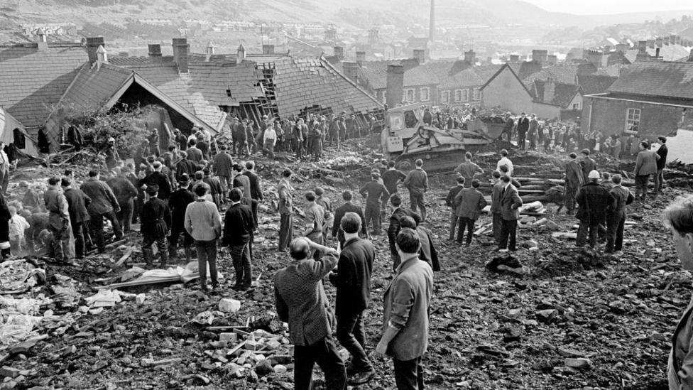 The Aberfan disaster - crowds of men look on in this black and white photograph standing on the rubble with a digger visible in the background