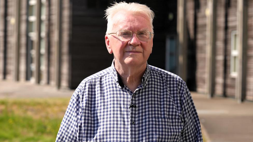A man with white hair and glasses standing on grass in front of a wooden school building