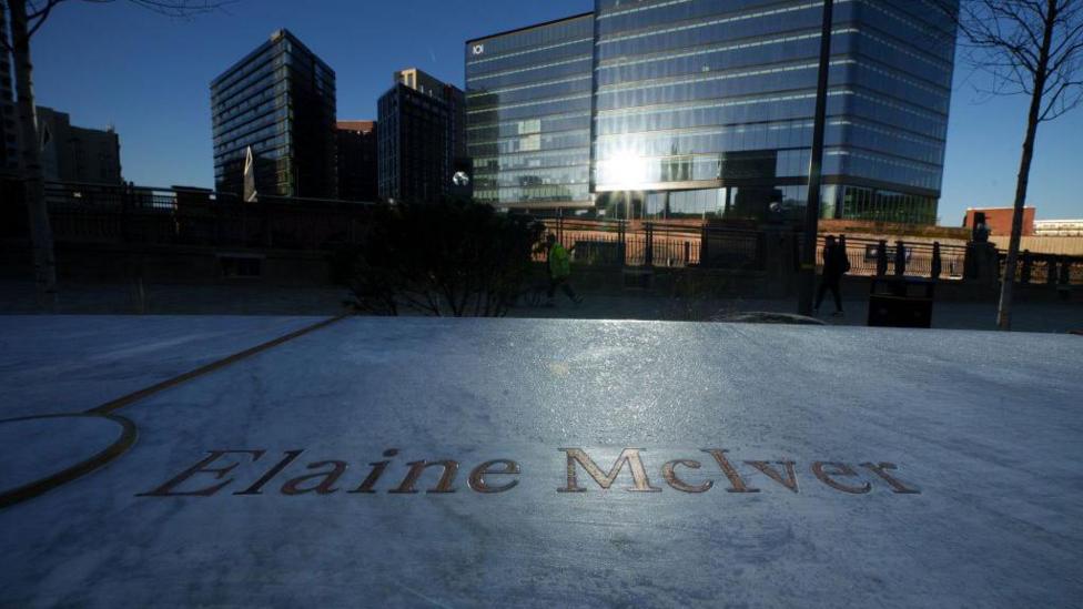 The name of Elaine McIver is engraved in gold on a marble slab, with buildings and the glinting sun in the background