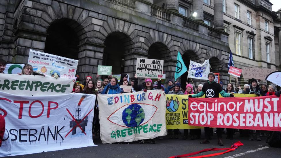 A group of protesters from climate organisations outside the Court of Session in Edinburgh. From left to right, one group are holding a greenpeace banner, which has the name of the organisation in green on a white banner. Below that is the words