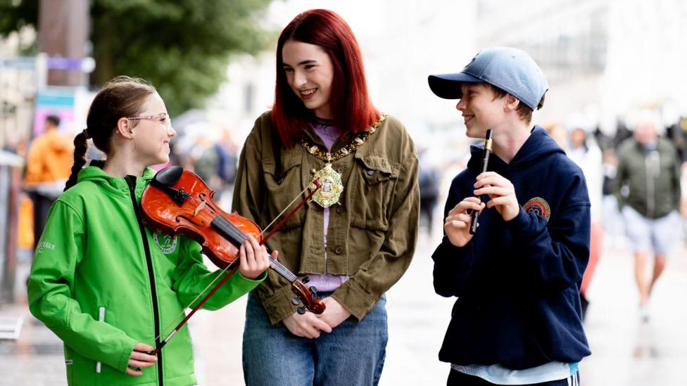 A girls in green jacket holding violin, a woman with dark green jacket, mayoral chains and denim jeans and a boy with navy jumper and blue hat holding a flute
