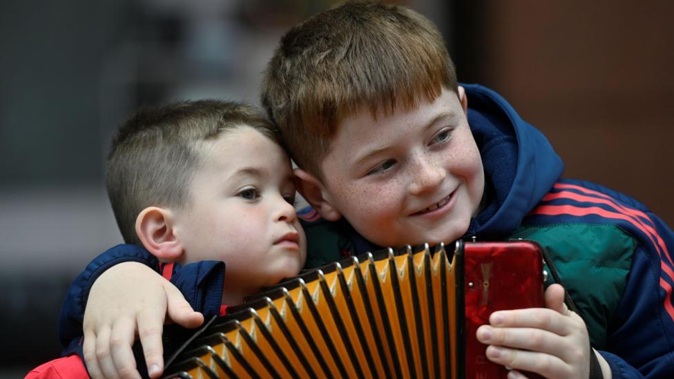 Two boys with dark hair hold an accordion and embrace