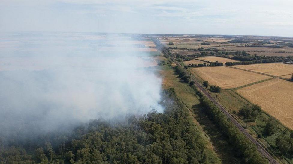 Smoke can be seen rising from a wooded area and there are farmers' fields in the distance