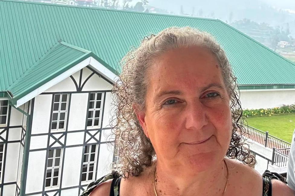 A woman in her 60s with grey curly hair pulled back from her face stands in front and slightly above of a large white building with black window frames and a green roof. She is wearing and has tie straps on her shoulders. In the distance a misty hill side is in view.