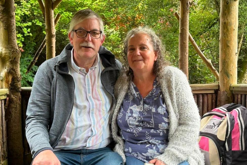 A man and a woman in their 60s sit in an open sided tree house where the trunks of the trees behind them are shown to hold up the platform. There is foliage in the background. The couple are sitting close together and are both wearing jeans. The man has glasses and a pastel striped shirt and is wearing an unzipped grey fleece over it. The woman has a blue top with white flowers with a grey fluffy cardigan over it. There is a black, pink and beige rucksack next to her.