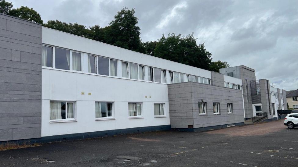 A grey and white two-storey building with a car park at the front
