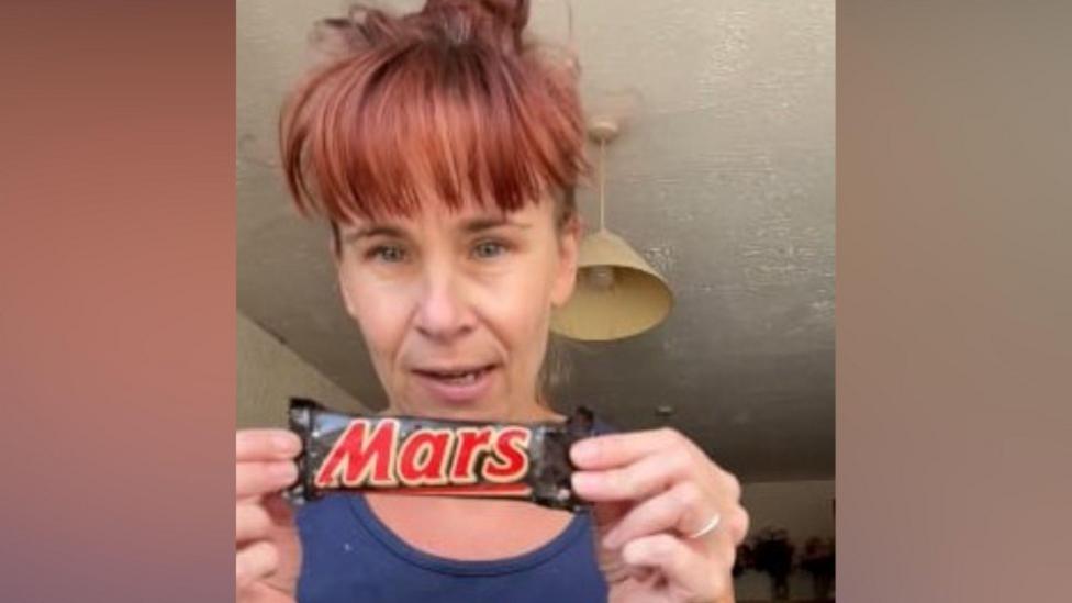 A woman holding up the old Mars Bar. She is wearing a navy vest and has red hair tied up with a full fringe.