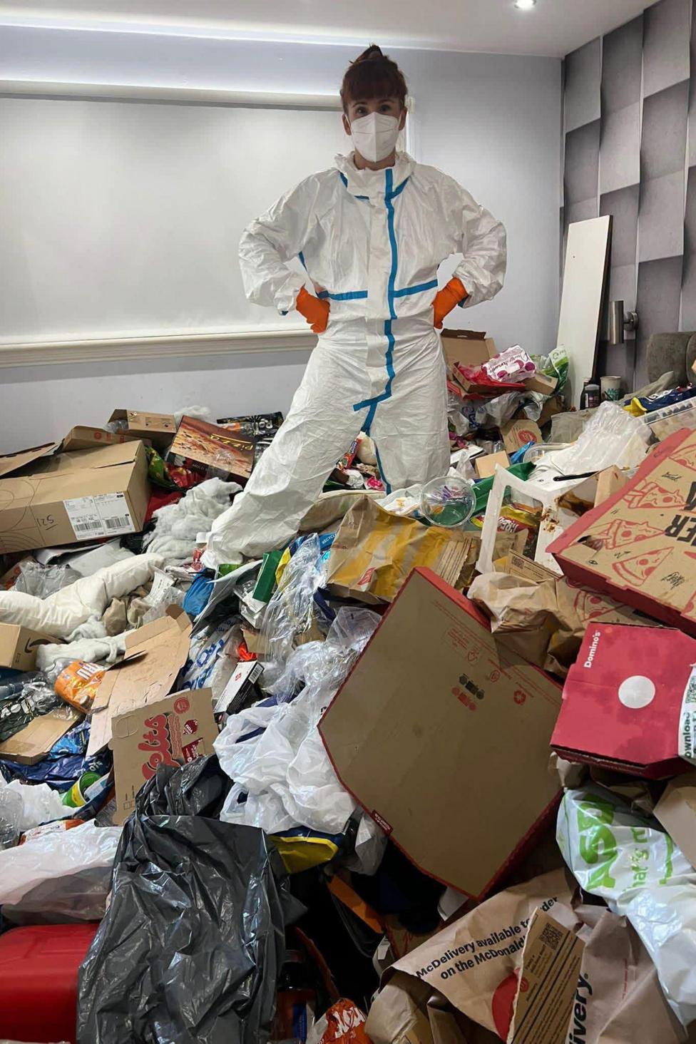 A woman in white overalls and orange gloves as well as a white mask is standing in a room full of cardboard and rubbish. She has her hands on her hips and her dark hair is tied up.