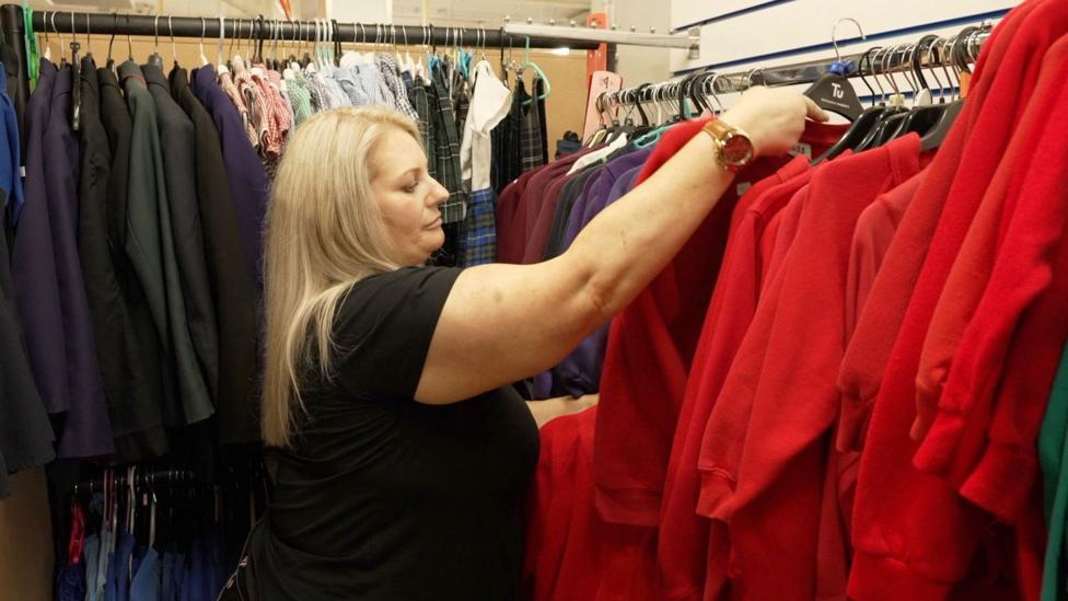 Michaela Wilson stands between two tightly packed rows of school uniforms on clothing rails inside a storeroom. She is reaching up with one hand to sort through a row of red sweatshirts hanging on black hangers