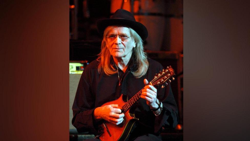 Henry McCullough Of Slim Chance, playing in 2004. He is dressed in black and holding a mandolin.He is wearing a black hat