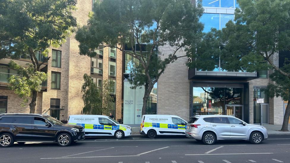 Two white Metropolitan Police Forensic Services vans parked along a street in front of a modern residential apartment building and office entrance displaying a 