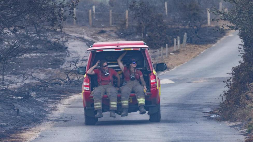 Two fire officers in the back of a fire service vehicle. The boot door is open and they are sitting in the back with their legs dangling from the vehicle. They are covered in smoke and wear sunglasses. The vehicle is moving down a road and either side is the blackened remains of trees and shrubbery.