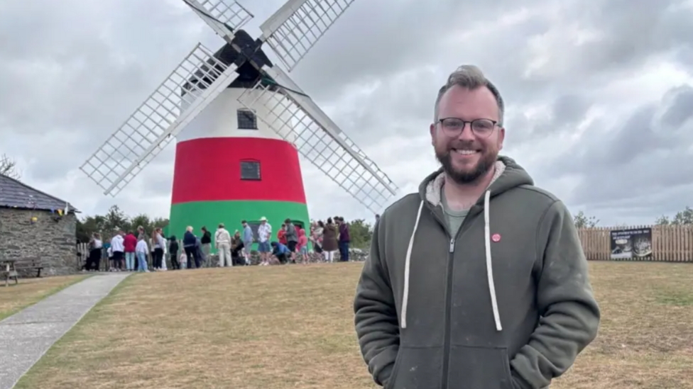 Richard Holt in front of a windmill painted red, white and green with people outside it. He has short blond hair and wears glasses.