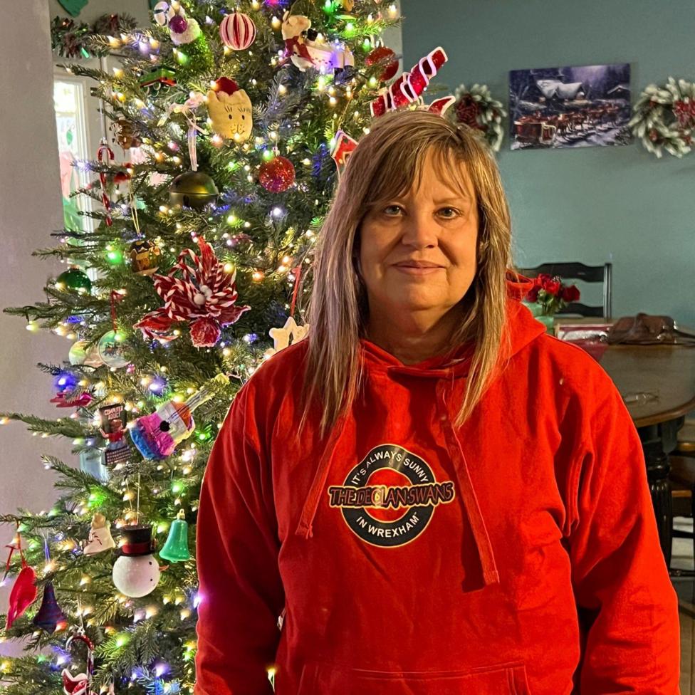 Janice in a red hoodie with a logo reading It's always sunny in Wrexham: The Declan Swans. She has blonde shoulder length hair and a fringe and is standing in front of a Christmas tree with white lights.