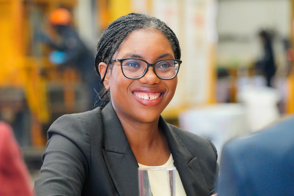 Kemi Badenoch smiling: she is visiting a factory and has her braided hair tied back. She wears glasses and a black jacket over a white blouse.