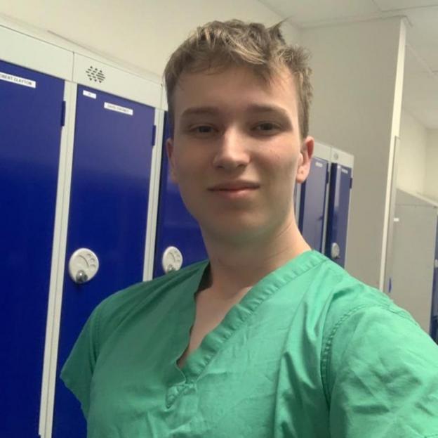A selfie of Harrison dressed in scrubs. The photo is taken in the changing room, the lockers are seen on the background.