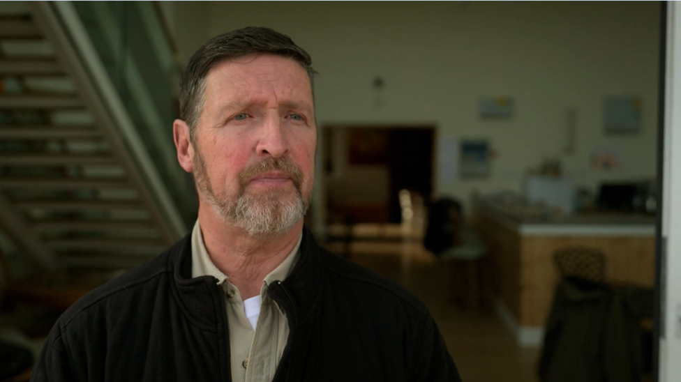 A waist-up portrait shows Michael inside a contemporary building. The individual wears a dark zip-up jacket over a light shirt. Behind him, an open-plan interior includes a staircase, seating areas and wooden furnishings, all softly out of focus to keep attention on the subject.
