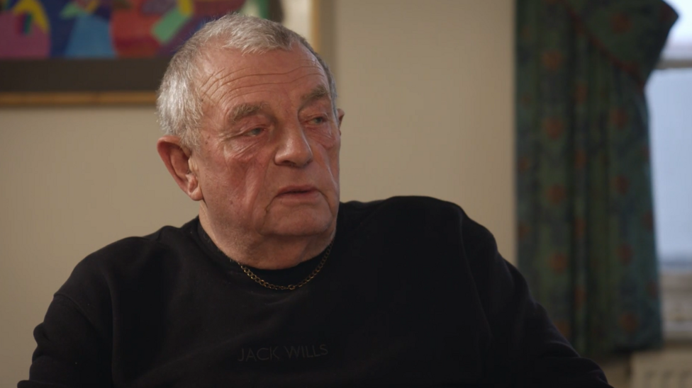 Paul Pemberton is photographed from the chest up inside a home or community setting. He has grey hair and wears a dark pullover and a thin chain necklace. The softly lit room includes a framed artwork on the wall and patterned curtains hanging beside a window, with the background slightly out of focus.
