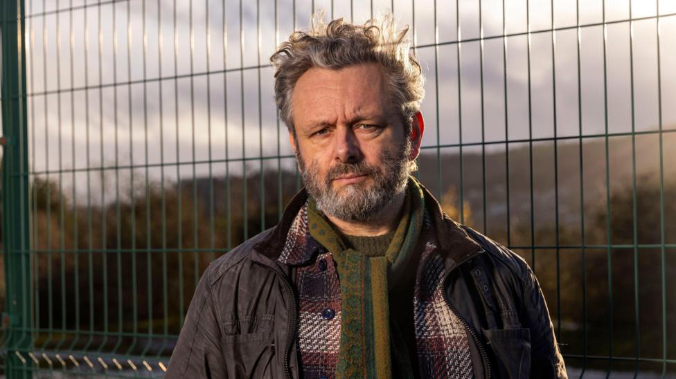 The image is a close outdoor portrait taken near a wire-mesh security fence. Michael Sheen stands facing the camera, dressed for cool weather in several layers, including a dark field jacket, a checked wool coat and a patterned green scarf. The fence fills much of the background, while distant hills and trees are softened by shallow depth of field. Golden sunlight from a low angle adds warmth to the scene and emphasises the textures of the scarf, coat and fence.