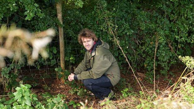 A person in an olive-green outdoor jacket crouches beside a wire fence at the edge of dense woodland. The ground is covered with leaves, grass and low vegetation, while thick green foliage fills the background. Softly blurred plants frame the foreground, drawing attention to the secluded countryside setting.