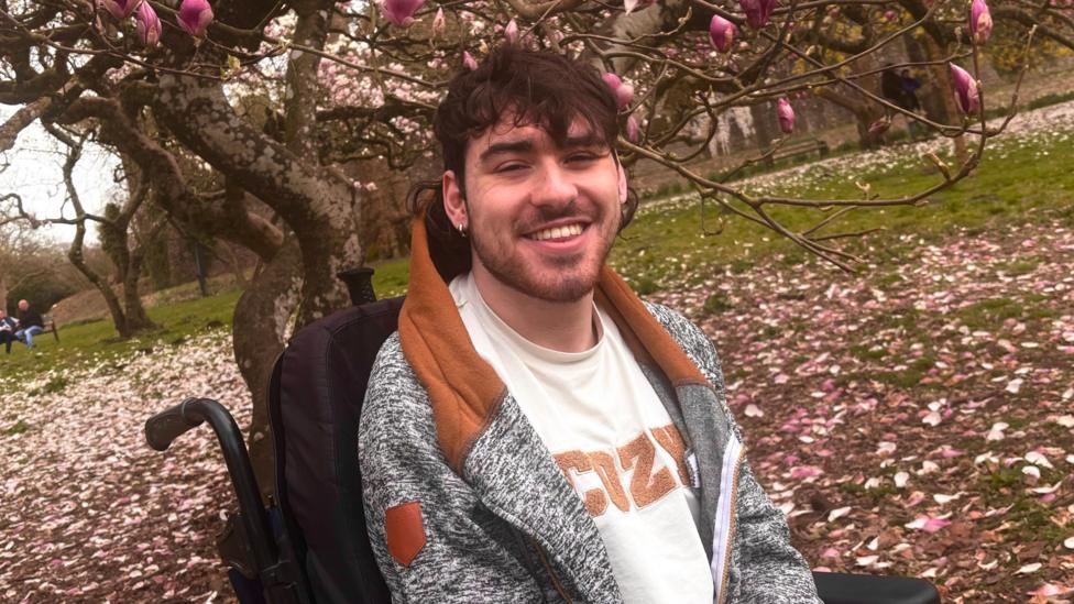 A smiling young man is seated outdoors in a park beneath blooming trees with pink flowers and fallen petals covering the ground. He has dark wavy hair and a light beard, and is wearing a gray and brown hoodie over a white shirt that says cozy, the backrest of his wheelchair is visible behind him.