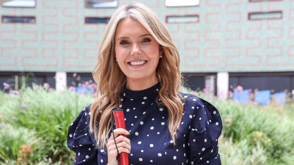 Smiling blonde woman with mid length wavy hair wearing a navy dress with white polka dots and long sleeves. She's holding a red-handled cane and standing outdoors in front of a patterned building with greenery and flowers in the background.
