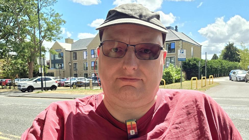 A close-up outdoor selfie of a man wearing a green cap, rectangular tinted glasses, and a maroon T-shirt. He is centered in the frame with a neutral expression, on a sunny street with parked cars, apartment buildings, trees, and a bright blue sky with clouds in the background.