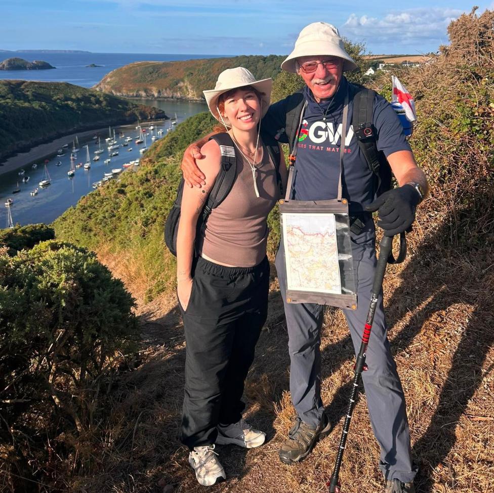 John Parry and Kyla Norrie stand smiling at the camera. They are both wearing white sun hats, and have their arm around each other's shoulders. John is wearing a 'Grandad's Great Walk' charity t-shirt, while Kyla wears a brown vest. The pair are standing on a footpath in north Pembrokeshire, with a view of hills behind them.