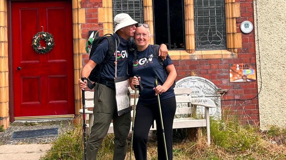 John kisses Hayley on the forehead, with his hand around her shoulder. They hold walking sticks, and stand in front of a red brick house with a red door.
