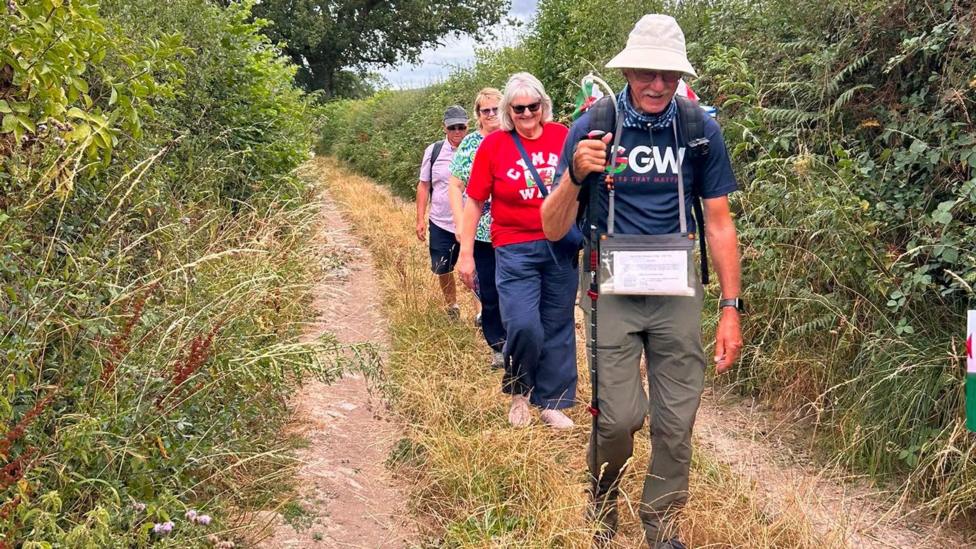 John leads the way down a narrow country lane, with tall hedges either side. He holds a walking stick, with his family members walking in a line behind him.