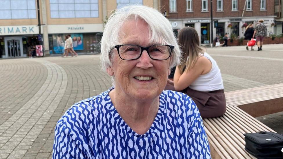 Barbara Logan is sitting on a bench in a square in Truro. She is wearing a blue and white patterned top and a pair of glasses. She has short white hair and is smiling. A woman with long dark blonde hair is sitting behind her. There are a few people walking across the square in the background and there is a Primark, a JD Sports and a Wetherspoons.
