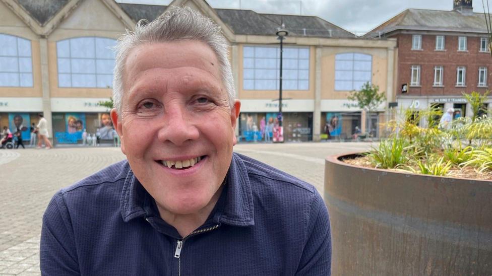 Nigel Brown is sitting on a bench in a square in Truro. He is wearing a blue T-shirt with a half-zip. He has short grey hair and is smiling broadly. To his left is a large concrete planter which has grasses in it. There are a few people walking across the square in the background and there is a Primark, a JD Sports and a Wetherspoons.