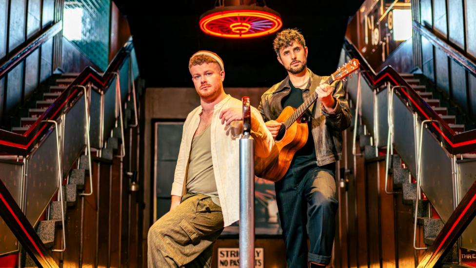 Two men, dressed casually, on a stage set showing twin flights of stairs. The man on the right has an acoustic guitar.