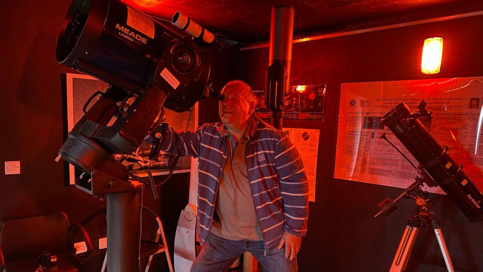 Publican Alan Boddington is wearing a blue and white striped jacket and looking through a very large telescope. There is a smaller telescope to his right.