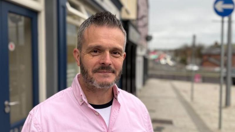 A shot of Robbie Butler looking down the lens of the camera. He's wearing a pink shirt and has grey hair and a greying beard.