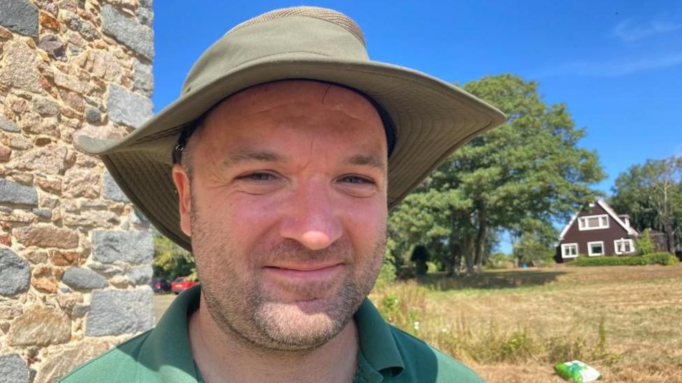 Will Harford-Fox is a man in his 30s He smiles at the camera and is wearing a khaki green broad brim hat and a bright green shirt. Behind him is a dry field, and a brown farmhouse with white window frames.