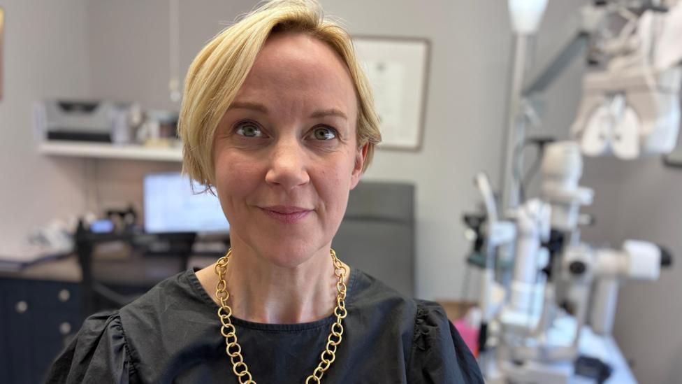 A woman with blonde hair in a short bob. She is wearing a black top and a gold chain necklace. She is in a room in an optician surgery. 