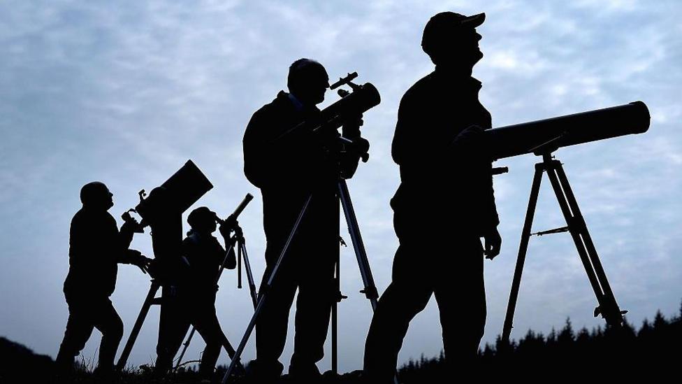 Silhouettes of astronomers setting up telescopes and cameras ahead of a solar eclipse. The sky is clouded over.