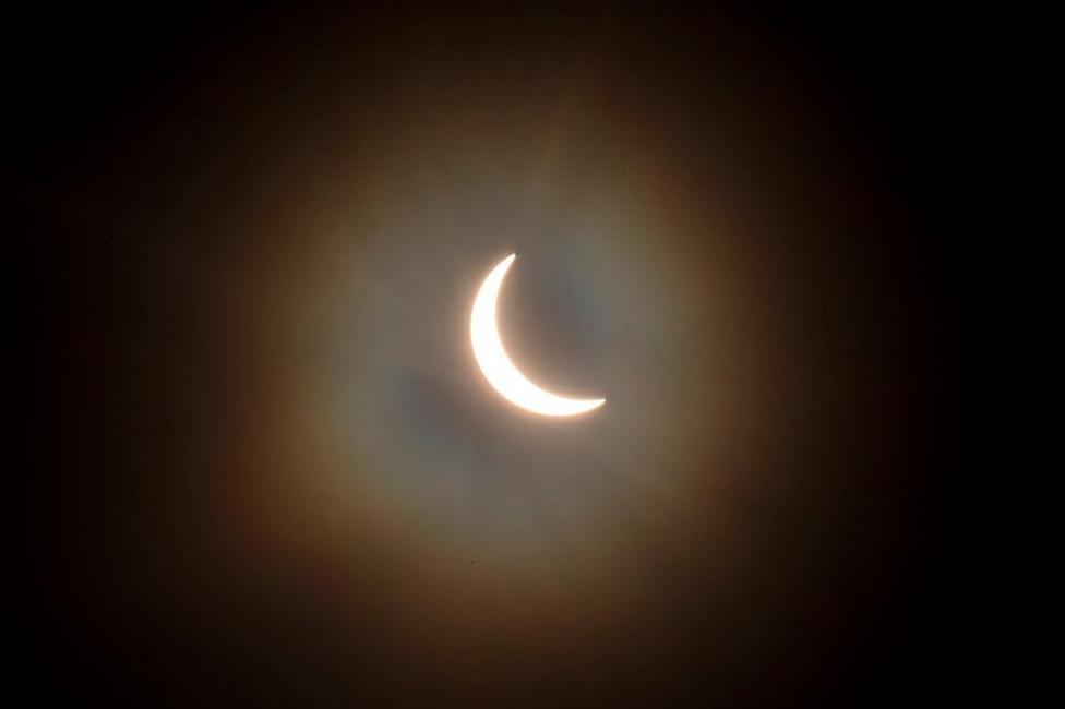 The sun is pictured during a partial solar eclipse. The sky is dark and cloudy. The sun is a thin arc in the sky.