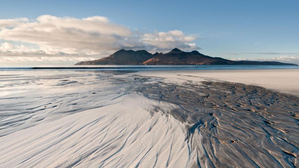 Rum is an island of red-coloured hills and viewed across a wide sandy beach on a sunny day. There are marks in the sand left by a falling tide.
