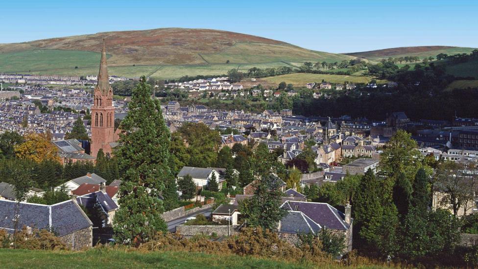 A general view of a town surrounded by green hills. There is a church steeple in the left of the picture.