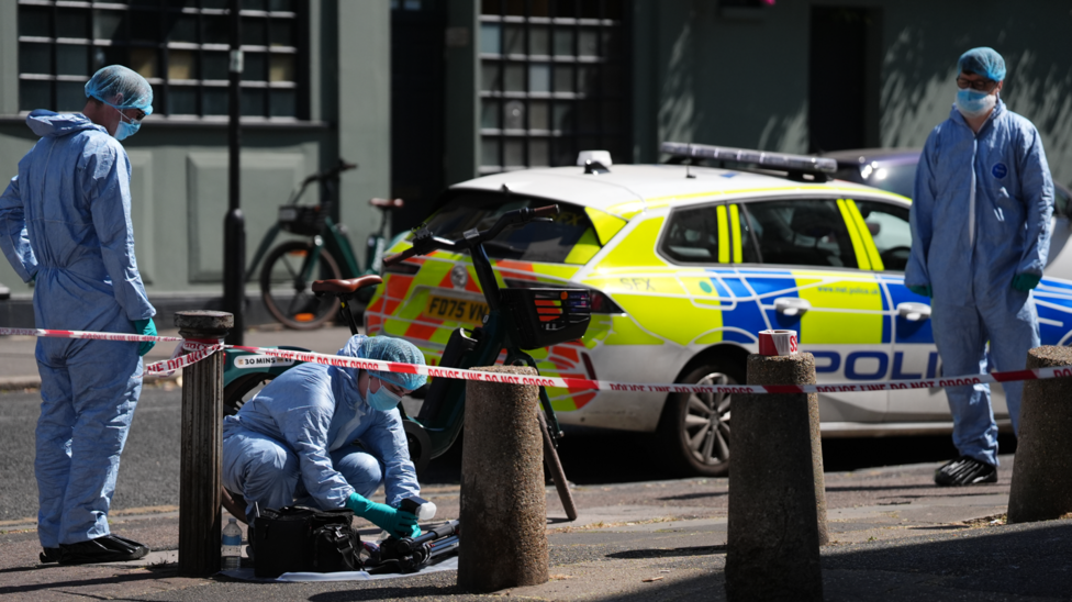 Forensic investigators in protective clothing examine a cordoned-off street scene near a bicycle, with police tape and a police car in the background.