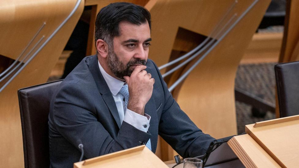 Humza Yousaf, dressed in a grey suit and light blue tie, sitting behind a desk in a debating chamber at Holyrood.