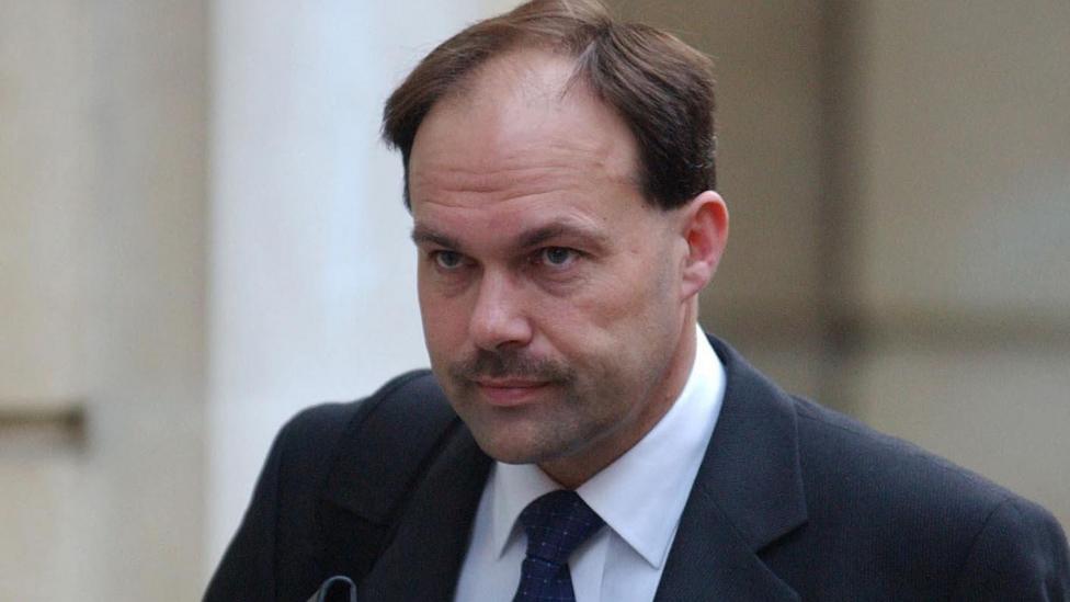 Glyn outside court during his 2003 trial. He has a grim look on his face, and has short brown hair and a moustache. He wears a black suit jacket and a dark tie, with a white shirt.