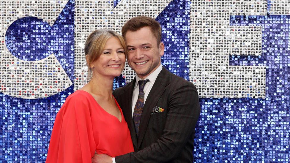An older blonde woman in a red dress and Egerton with brown hair and wearing a suit. They're on a blue