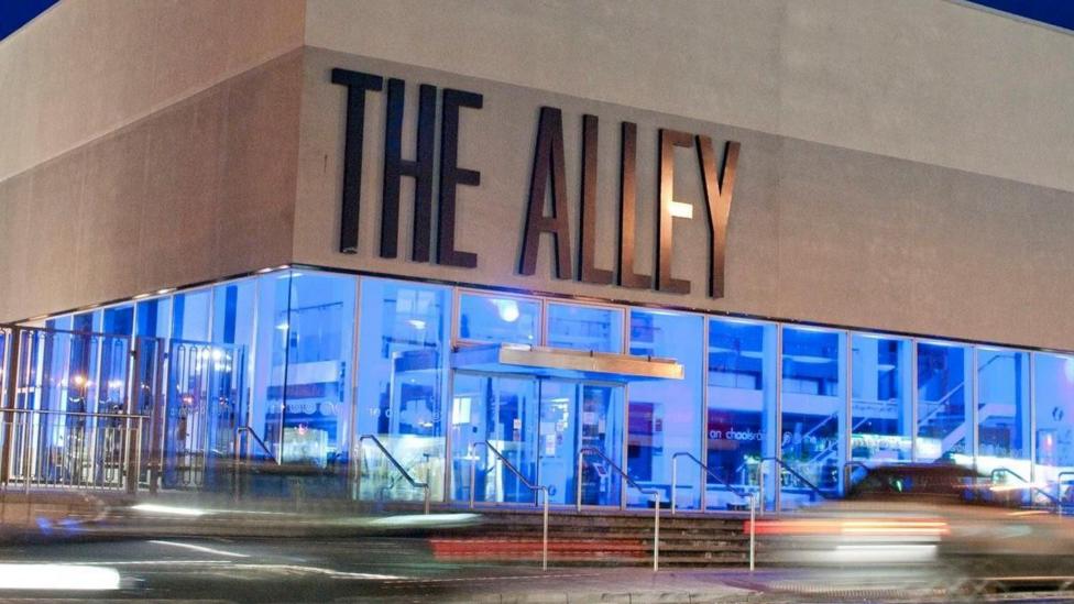 The Alley Theatre in Strabane. The theatre has its name on the front in large grey letters. The building is grey with glass panels around it on all sides. The lights inside are blue. The forefront of the picture is blurry. There are steps leading to the door of the theatre. Copyright