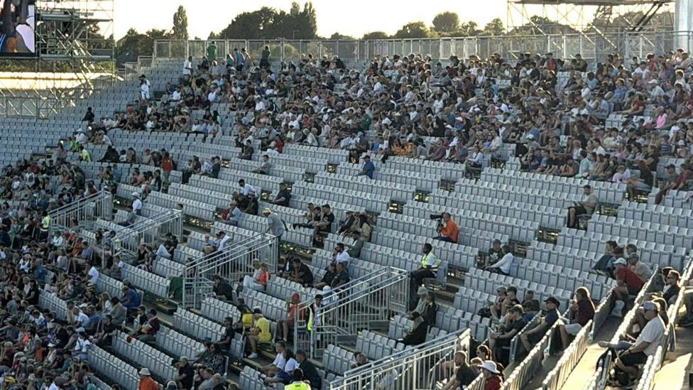 A photo of stands at a stadium. There are empty seats which are grey. You can also see fans sitting in different rows.