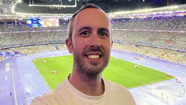A photo of a man smiling in front of a stadium. The stadium is purple and the man has short brown hair and has a beard. He is wearing a while T-shirt.